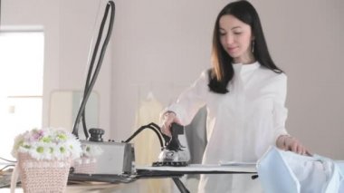 Genç kızın yavaş videosu ütülüyor. Elbiseleri askıya asıyor, dumanlar dikey durumda, buharlı profesyonel bir ütü, dikiş üretimi, dikiş stüdyosu, ajans...