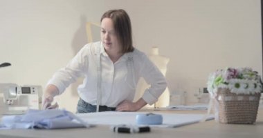 Moda tasarımcısının yavaş çekim videosu kıvrımlı ve desenli bir masada çalışıyor, kumaşı kesiyor, etrafında makas, santimetre, dikiş makinesi ve arka planda kukla, pürüzsüz bir hareket var.