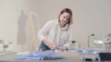Moda tasarımcısının yavaş çekim videosu kıvrımlı ve desenli bir masada çalışıyor, kumaşı kesiyor, etrafında makas, santimetre, dikiş makinesi ve arka planda kukla, pürüzsüz bir hareket var.