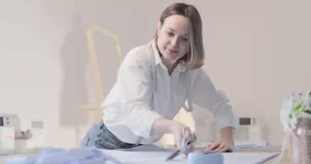 Повільне відео модного дизайнера працює за столом з кривими і візерунком, вона ріже тканину, навколо лежить ножиці, сантиметр, швейна машина і манекен на фоні, плавний рух — стокове відео