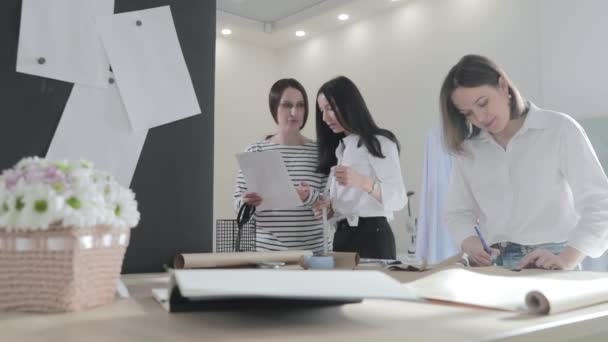 Повільне відео з трьох дизайнерів моди працює за столом і обговорює щось, вигини і візерунок, папір для ножиць, дівчат, які дивляться в смартфон, шиють студію, сонячне світло, посміхаються — стокове відео