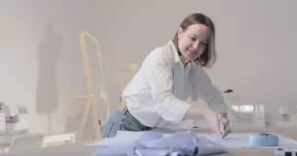 Zpomalené video módní návrhářka pracuje u stolu s křivkami a vzor, ona řezání tkaniny, kolem leží nůžky, centimetr, šicí stroj a figuríny na pozadí, Hladký pohyb — Stock video