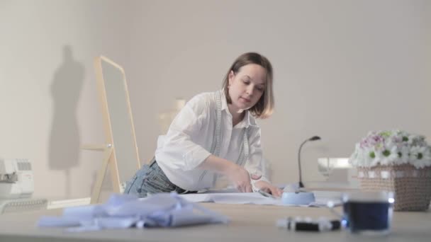 Повільне відео модного дизайнера працює за столом з кривими і візерунком, вона ріже тканину, навколо лежить ножиці, сантиметр, швейна машина і манекен на фоні, плавний рух — стокове відео