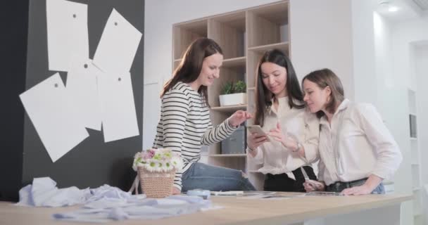 Vídeo en cámara lenta de los tres diseñadores de moda trabaja en una mesa y discutir sobre algo, curvas y un patrón, papel de tijeras, niñas arelooking en el teléfono inteligente, estudio de costura, luz solar, sonrisas — Vídeo de stock