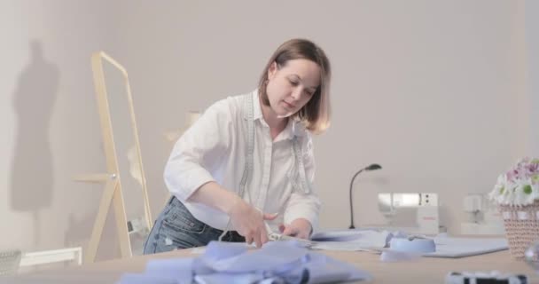 Zeitlupenvideo der Modedesignerin arbeitet an einem Tisch mit Kurven und einem Muster, sie schneidet Stoff, um sie herum liegt Schere, Zentimeter, Nähmaschine und Schnuller auf dem Hintergrund, sanfte Bewegung — Stockvideo