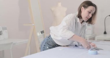 Moda tasarımcısının yavaş çekim videosu kıvrımlı ve desenli bir masada çalışıyor, kumaşı kesiyor, etrafında makas, santimetre, dikiş makinesi ve arka planda kukla, pürüzsüz bir hareket var.