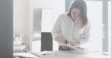 Moda tasarımcısının yavaş çekim videosu kıvrımlı ve desenli bir masada çalışıyor, kumaşı kesiyor, etrafında makas, santimetre, dikiş makinesi ve arka planda kukla, pürüzsüz bir hareket var.