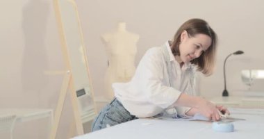 Moda tasarımcısının yavaş çekim videosu kıvrımlı ve desenli bir masada çalışıyor, kumaşı kesiyor, etrafında makas, santimetre, dikiş makinesi ve arka planda kukla, pürüzsüz bir hareket var.