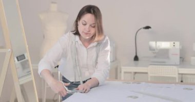 Moda tasarımcısının yavaş çekim videosu kıvrımlı ve desenli bir masada çalışıyor, kumaşı kesiyor, etrafında makas, santimetre, dikiş makinesi ve arka planda kukla, pürüzsüz bir hareket var.