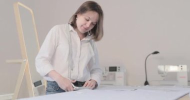 Moda tasarımcısının yavaş çekim videosu kıvrımlı ve desenli bir masada çalışıyor, kumaşı kesiyor, etrafında makas, santimetre, dikiş makinesi ve arka planda kukla, pürüzsüz bir hareket var.