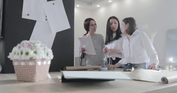 Zeitlupenvideo der drei Modedesignerinnen arbeitet an einem Tisch und diskutiert über etwas, Kurven und Muster, Scherenpapier, Mädchen, die im Smartphone herumlungern, Nähstudio, Sonnenlicht, Lächeln — Stockvideo