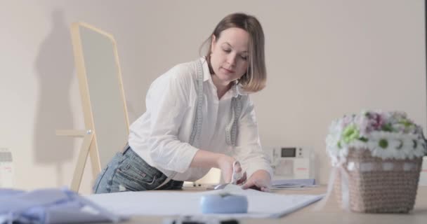 Vidéo au ralenti du créateur de mode travaille à une table avec des courbes et un motif, elle coupe le tissu, autour des mensonges ciseaux, centimètre, machine à coudre et mannequin sur le fond, mouvement lisse — Video