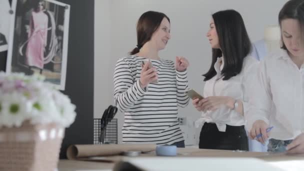 Rusia, San Petersburgo, 01 Marzo 2019: Tres trabajos de diseñador de moda en el estudio de costura, las niñas discuten sobre algo y se ve en el teléfono inteligente, una mesa con curvas y un patrón, Sala de luz, cámara lenta — Vídeos de Stock