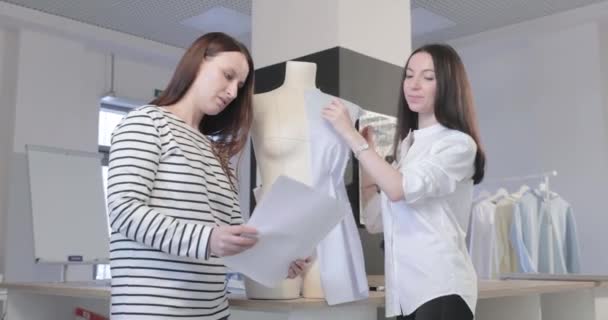 Το βίντεο αργής κίνησης δύο κοριτσιών του σχεδιαστή μόδας συζητούν για κάτι, κοιτάζουν ένα φύλλο χαρτί στο στούντιο ραπτικής, το σκίτσο μιας φωτογραφίας του μοντέλου στα χέρια, μια κούκλα στο φόντο — Αρχείο Βίντεο
