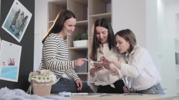 Rusya, St.Petersburg, 01 Mart 2019: Üç moda tasarımcısı dikiş stüdyosunda çalışıyor, kızlar bir şey hakkında tartışıyor ve akıllı telefondan bakıyor, kıvrımlı ve şablonlu bir masa, hafif çekimli, yavaş çekim — Stok video