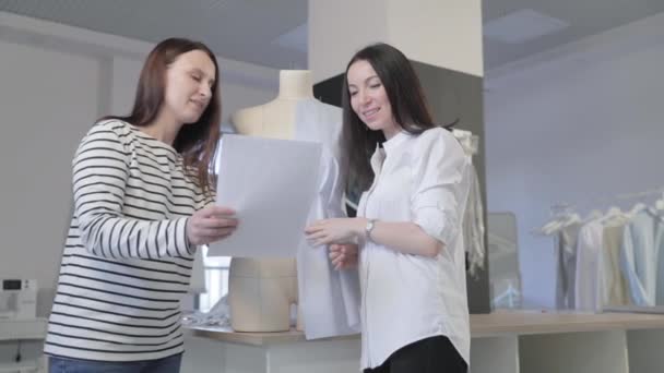 O vídeo de câmera lenta de duas meninas do desenhista de moda discute sobre algo, está olhando para uma folha de papel no estúdio de costura, o esboço de uma foto do modelo em mãos, um manequim no fundo — Vídeo de Stock