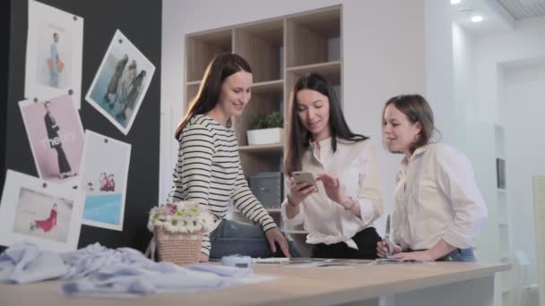 Rosja, Sankt Petersburg, 01 marzec 2019: Trzy projektantki mody pracują w studio szycia, dziewczyny o czymś dyskutują i patrzą na smartfona, stół z krzywiznami i wzorem, Sala świetlna, zwolnione tempo — Wideo stockowe