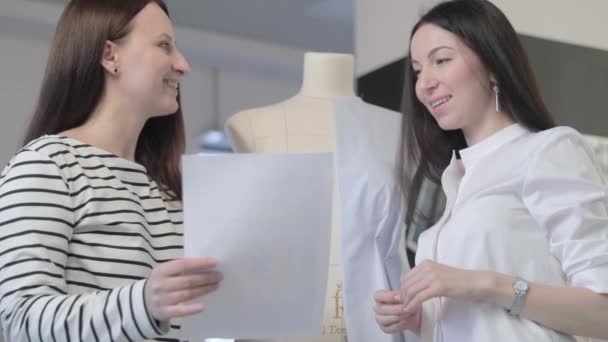 Vídeo en cámara lenta de dos chicas del diseñador de moda discutir sobre algo, están mirando una hoja de papel en el estudio de costura, el boceto de una foto de modelo en las manos, un maniquí en el fondo — Vídeo de stock