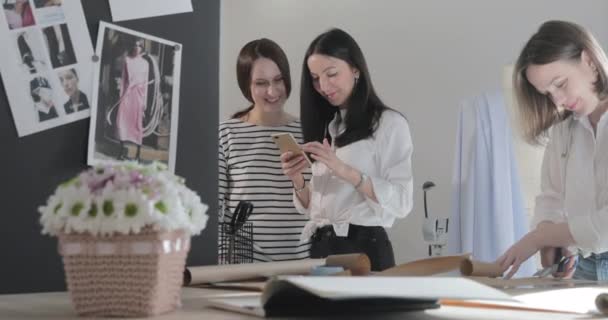 Russie, Saint-Pétersbourg, 01 mars 2019 : Trois créateurs de mode travaillent au studio de couture, les filles discutent de quelque chose et regarde dans le smartphone, une table avec des courbes et un motif, salle de lumière, ralenti — Video