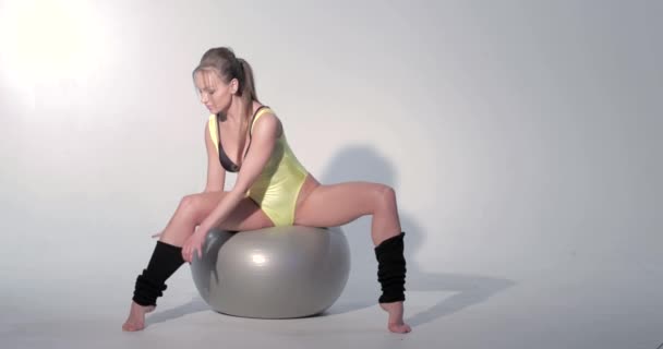 Beautiful girl in a yellow sports suit and black leggings is engaged in gymnastics on a gray ball, she flirts, barefoot, long hair in the tail, Extension on the ball, is jumping, Black gaiters — Stock Video