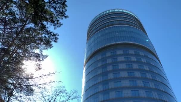 Rusko, Petrohrad, 7. května 2020: Národní centrum lékařského výzkumu VA Almazov, pohled přes větve stromů se zelenými listy, modré nebe na pozadí, odrazy slunce na skle fasády — Stock video