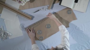 Masada kıvrımlı ve desenli çalışan moda tasarımcısının yavaş çekim videosu, bir sabun dilimi çizer, etrafında makas, santimetre, çizimli bir defter, dikiş stüdyosu, üst kattan görünüm