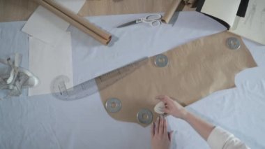 Masada kıvrımlı ve desenli çalışan moda tasarımcısının yavaş çekim videosu, bir sabun dilimi çizer, etrafında makas, santimetre, çizimli bir defter, dikiş stüdyosu, üst kattan görünüm