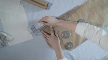 Masada kıvrımlı ve desenli çalışan moda tasarımcısının yavaş çekim videosu, bir sabun dilimi çizer, etrafında makas, santimetre, çizimli bir defter, dikiş stüdyosu, üst kattan görünüm