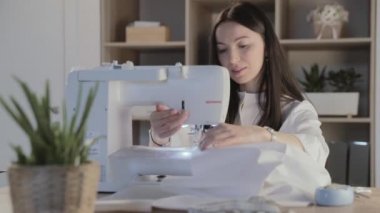 Rusya, Saint-Petersburg, 01 Mart 2019: Güzel genç kız dikiş atölyesindeki otomatik dikiş makinesinde oturuyor ve bazı kıyafetler dikiyor, esmer, uzun saçlı, çiçekli ve aksesuarlı.