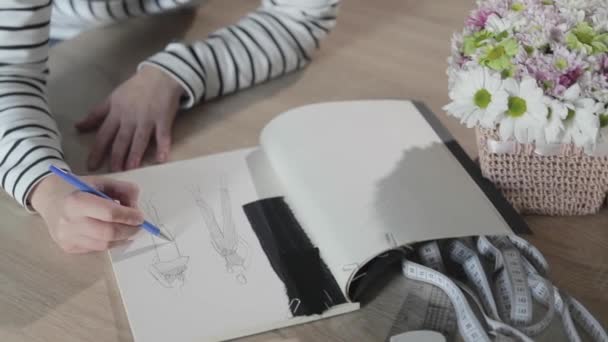 Vidéo au ralenti de la jeune fille dessine le croquis du futur modèle de vêtements dans le carnet, outils pour la couture, agence de couture, un bouquet de fleurs, artisanat en papier, vue de dessus — Video