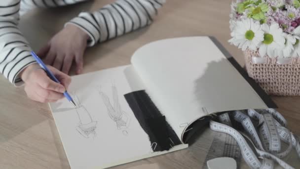 Vidéo au ralenti de la jeune fille dessine le croquis du futur modèle de vêtements dans le carnet, outils pour la couture, agence de couture, un bouquet de fleurs, artisanat en papier, vue de dessus — Video