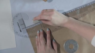 Masada kıvrımlı ve desenli çalışan moda tasarımcısının yavaş çekim videosu, bir sabun dilimi çizer, etrafında makas, santimetre, çizimli bir defter, dikiş stüdyosu, üst kattan görünüm