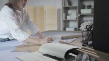 Masada kıvrımlı ve desenli çalışan moda tasarımcısının yavaş çekim videosu, bir sabun dilimi çizer, etrafında makas, santimetre, çizimli bir defter, dikiş stüdyosu, üst kattan görünüm