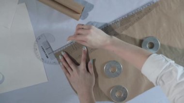 Masada kıvrımlı ve desenli çalışan moda tasarımcısının yavaş çekim videosu, bir sabun dilimi çizer, etrafında makas, santimetre, çizimli bir defter, dikiş stüdyosu, üst kattan görünüm