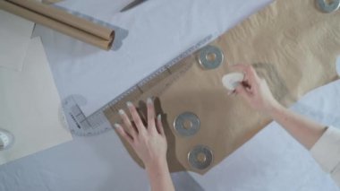 Masada kıvrımlı ve desenli çalışan moda tasarımcısının yavaş çekim videosu, bir sabun dilimi çizer, etrafında makas, santimetre, çizimli bir defter, dikiş stüdyosu, üst kattan görünüm