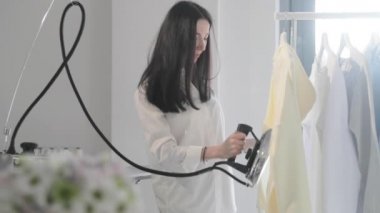 Genç kızın yavaş çekim videosu, korkuluklara asılı giysileri ütülüyor, dikey vaziyette buharlar çıkıyor, buharlı profesyonel bir ütü, dikiş üretimi, stüdyo, ajans...