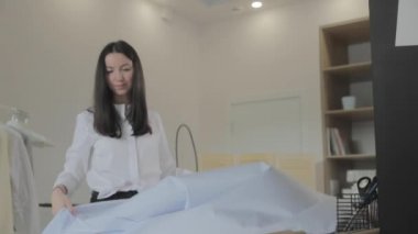 Genç, uzun saçlı esmer kız dikiş atölyesinde terzilik için bir masaya kumaş seriyor, mavi renk, el işi kağıt.
