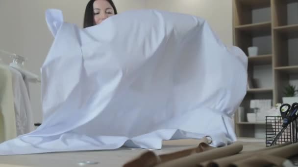 De jonge langharige brunette meisje spreidt stof op een tafel voor het maken van jurken in naaisalon, blauwe kleur, ambachtelijk papier — Stockvideo