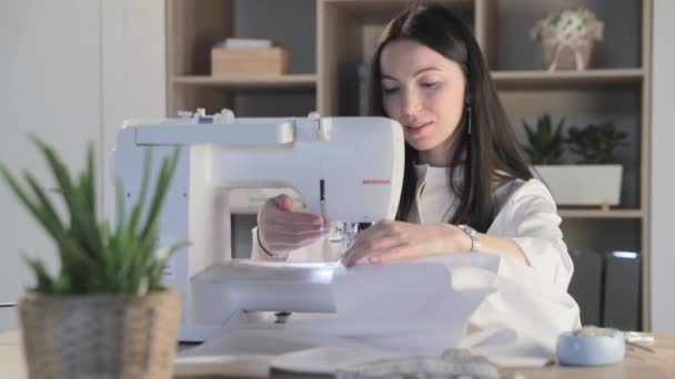 Russia, Saint-Petersburg, 01 March 2019: The beautiful young girl sits at the automatic sewing machine in sewing studio and sews some clothes, the brunette, long hair, flowers and accessories — Stock Video