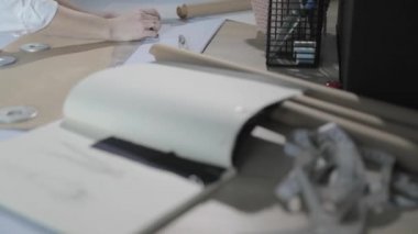 Masada kıvrımlı ve desenli çalışan moda tasarımcısının yavaş çekim videosu, bir sabun dilimi çizer, etrafında makas, santimetre, çizimli bir defter, dikiş stüdyosu, üst kattan görünüm