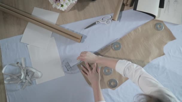 Slow motion video van de mode-ontwerper die werkt aan de tafel met bochten en een patroon, tekent een stuk zeep, rond ligt een schaar, centimeter, een notitieboekje met schets, naaiatelier, uitzicht vanaf een top — Stockvideo