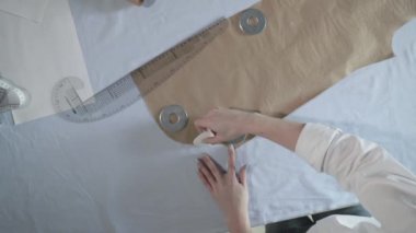 Masada kıvrımlı ve desenli çalışan moda tasarımcısının yavaş çekim videosu, bir sabun dilimi çizer, etrafında makas, santimetre, çizimli bir defter, dikiş stüdyosu, üst kattan görünüm