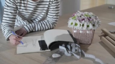 Kızın yavaş çekim videosu not defterine gelecekteki kıyafet modelinin taslağını çizer, dikiş, dikiş ajansı, bir demet çiçek, kağıt işçiliği, üst görünüm