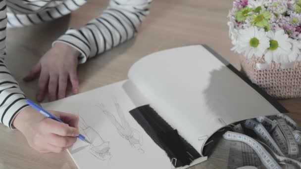 Відео повільного руху дівчини малює ескіз майбутньої моделі одягу в блокноті, інструменти для швейного, швейного агентства, купу квітів, паперових виробів, вид зверху — стокове відео