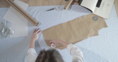 Masada kıvrımlı ve desenli çalışan moda tasarımcısının yavaş çekim videosu, bir sabun dilimi çizer, etrafında makas, santimetre, çizimli bir defter, dikiş stüdyosu, üst kattan görünüm