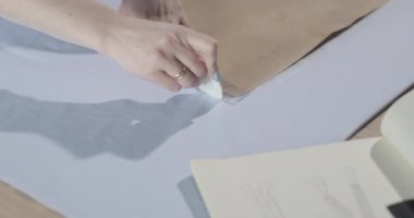 Masada kıvrımlı ve desenli çalışan moda tasarımcısının yavaş çekim videosu, bir sabun dilimi çizer, etrafında makas, santimetre, çizimli bir defter, dikiş stüdyosu, üst kattan görünüm