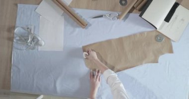 Masada kıvrımlı ve desenli çalışan moda tasarımcısının yavaş çekim videosu, bir sabun dilimi çizer, etrafında makas, santimetre, çizimli bir defter, dikiş stüdyosu, üst kattan görünüm