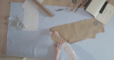 Masada kıvrımlı ve desenli çalışan moda tasarımcısının yavaş çekim videosu, bir sabun dilimi çizer, etrafında makas, santimetre, çizimli bir defter, dikiş stüdyosu, üst kattan görünüm