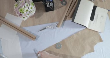 Masada kıvrımlı ve desenli çalışan moda tasarımcısının yavaş çekim videosu, bir sabun dilimi çizer, etrafında makas, santimetre, çizimli bir defter, dikiş stüdyosu, üst kattan görünüm