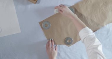 Masada kıvrımlı ve desenli çalışan moda tasarımcısının yavaş çekim videosu, bir sabun dilimi çizer, etrafında makas, santimetre, çizimli bir defter, dikiş stüdyosu, üst kattan görünüm